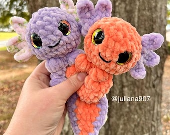 May include: Two handmade crochet axolotl plushies. One is purple with pink and green accents, the other is orange with purple accents. Both have large, iridescent eyes and smiling faces. The plushies are soft and cuddly, perfect for a child's toy or room decor.