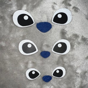 May include: Three sets of white eyes with black pupils and a blue nose on a gray background. The eyes and nose are felt appliques.