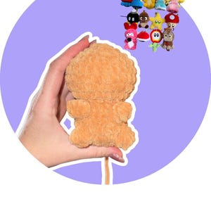 May include: A light brown, plush mushroom-shaped toy is held in a hand, with a collection of small, colourful crocheted character toys in the background. The mushroom toy has a soft, textured appearance. The background is a light purple circle.