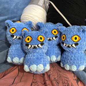 May include: Four blue crocheted monster plushies with large yellow and orange eyes, white fangs, and small feet. The plushies are arranged in a group, with white and black yarn in the background.