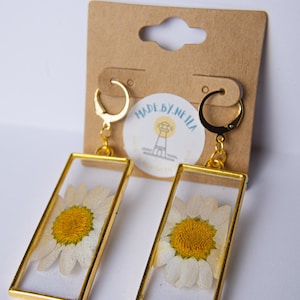 Könnte beinhalten: Goldene Creolen mit einem rechteckigen Rahmen, in dem eine gepresste weiße Margerite mit gelber Mitte zu sehen ist. Die Ohrringe befinden sich auf einer braunen Karte mit dem Text "MADE BY NEILA" und einer Leuchtturmgrafik.