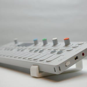 Könnte beinhalten: Ein hellgrauer OP-1 Synthesizer mit einem weißen Ständer. Das Gerät hat Knöpfe mit farbigen Oberteilen, darunter blau, grün und orange. Es verfügt über eine Tastatur und verschiedene Anschlüsse.
