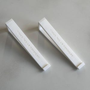May include: Two white, rectangular, 3D-printed objects with a handle and a flat base. The text "DESIGNED BY CRL CRLL" is printed on the side of each object. The objects are lying on a light grey surface.