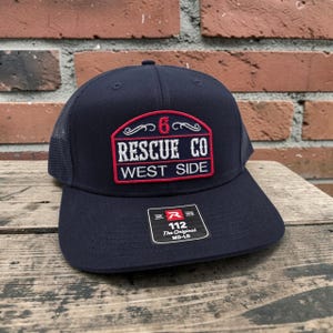 May include: A navy blue trucker hat with a mesh back and a structured front panel. An embroidered patch features the red number 6, the white text RESCUE CO, and WEST SIDE. A black sticker on the brim displays the text 112 The Original MD-LG.
