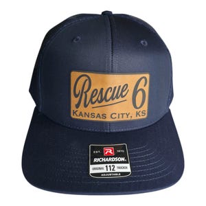 May include: Navy blue trucker hat with a brown leather patch. The patch features the text "Rescue 6 Kansas City, KS" in a script font. The hat has a curved brim and an adjustable strap.