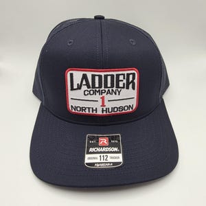 Op de afbeelding: Marineblauwe truckerpet met een mesh achterkant en een gebogen klep. De voorkant heeft de tekst "LADDER COMPANY 1 NORTH HUDSON" in witte en rode letters. Een klein label leest "RICHARDSON ORIGINAL 112 TRUCKER ADJUSTABLE".