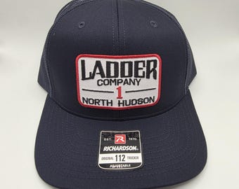 Könnte beinhalten: Marineblaue Trucker-Kappe mit Mesh-Rückseite und gebogenem Schirm. Der vordere Aufnäher zeigt "LADDER COMPANY 1 NORTH HUDSON" in weißer und roter Schrift. Ein kleines Etikett lautet "RICHARDSON ORIGINAL 112 TRUCKER ADJUSTABLE".