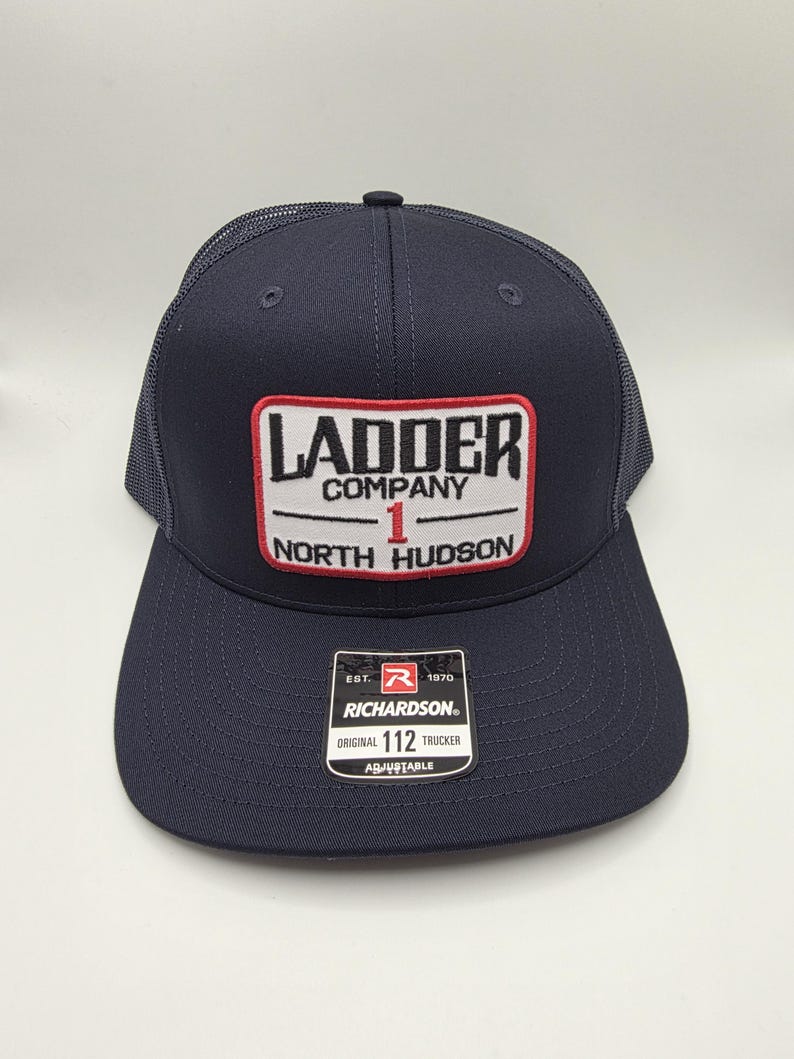 May include: Navy blue trucker hat with a mesh back and a curved brim. The front patch reads "LADDER COMPANY 1 NORTH HUDSON" in white and red lettering. A small tag reads "RICHARDSON ORIGINAL 112 TRUCKER ADJUSTABLE".