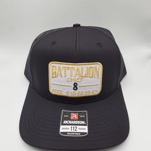May include: Black trucker hat with a mesh back and a curved brim. The front features a rectangular patch with "BATTALION CHIEF 8" embroidered in gold and white. The hat includes a "RICHARDSON" tag and is adjustable.