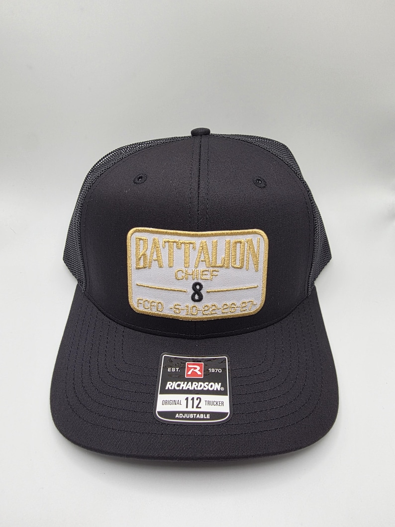 May include: Black trucker hat with a mesh back and a curved brim. The front features a rectangular patch with "BATTALION CHIEF 8" embroidered in gold and white. The hat includes a "RICHARDSON" tag and is adjustable.