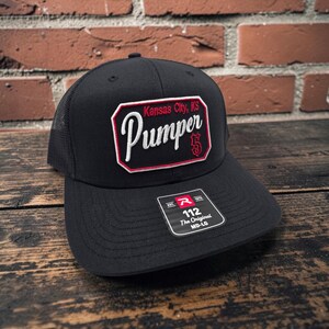 May include: A black trucker cap with a mesh back and a rectangular patch on the front. The patch features the text Kansas City, KS and Pumper 5 in white and red embroidery. A black sticker on the brim displays the text 112 The Original MD-LG.