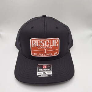 May include: Black trucker hat with a mesh back and a rectangular patch. The patch reads "RESCUE COMPANY 1 PHILADELPHIA, PA" in white and orange lettering. The hat also has a Richardson tag.