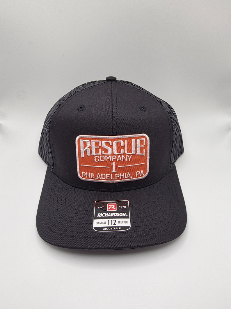 May include: Black trucker hat with a mesh back and a rectangular patch. The patch reads "RESCUE COMPANY 1 PHILADELPHIA, PA" in white and orange lettering. The hat also has a Richardson tag.