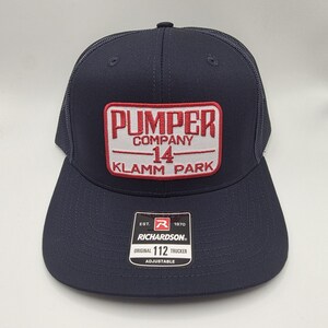 May include: Navy blue trucker hat with a rectangular patch featuring the text "PUMPER COMPANY 14 KLAMM PARK" in red and white. The hat has a curved brim and a mesh back. The Richardson brand label is visible.