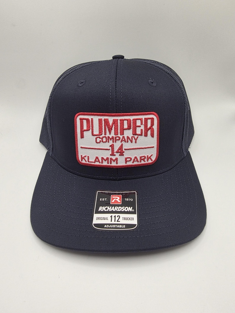 May include: Navy blue trucker hat with a rectangular patch featuring the text "PUMPER COMPANY 14 KLAMM PARK" in red and white. The hat has a curved brim and a mesh back. The Richardson brand label is visible.