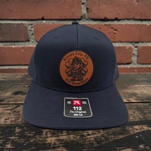 May include: A navy blue baseball cap featuring a circular brown leather patch on the front. The patch displays a graphic of a firefighter character and the text Kansas City, KS Fire Department. A black sticker on the brim reads 112 The Original MD-LG.