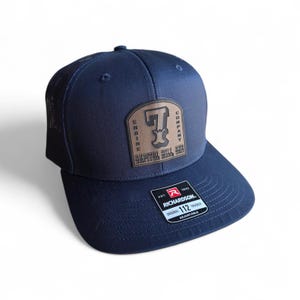 May include: A navy blue trucker cap with a brown leather patch displaying the text "ENGINE COMPANY 7 CAPITOL HILL, OKC". The cap has a black mesh back and a Richardson 112 label.