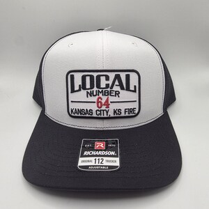 May include: A black and gray trucker hat with a white front panel. The hat features a patch with the text "LOCAL NUMBER 64 KANSAS CITY, KS FIRE." The brim is black, and the hat is adjustable.