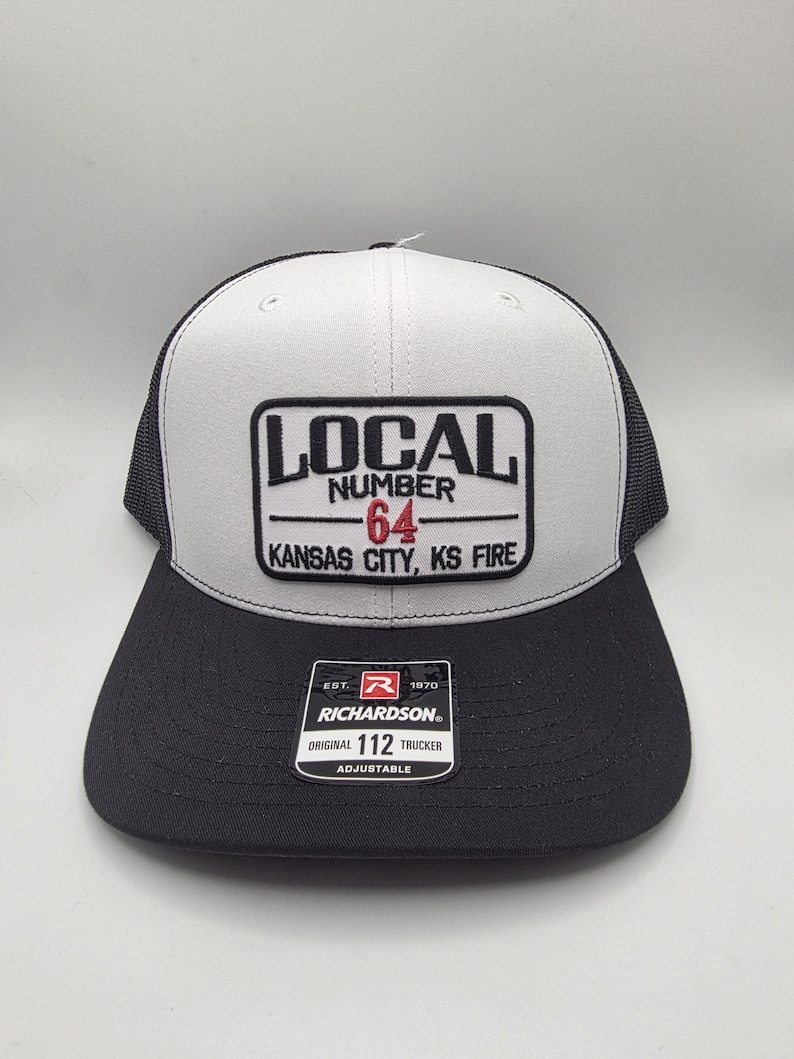 May include: A black and gray trucker hat with a white front panel. The hat features a patch with the text "LOCAL NUMBER 64 KANSAS CITY, KS FIRE." The brim is black, and the hat is adjustable.