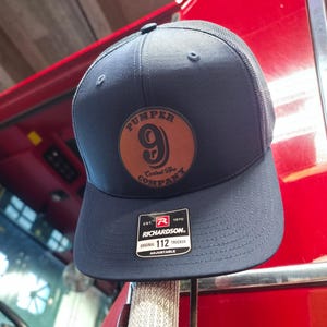 May include: A navy blue trucker cap with a brown leather patch displaying "PUMPER 9 Central Ave Company". The cap has a mesh back and a curved peak. The tag reads "RICHARDSON ORIGINAL 112 TRUCKER ADJUSTABLE".