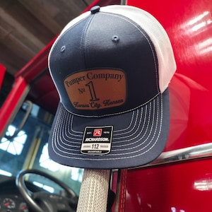 May include: Navy blue and white trucker hat with a brown leather patch. The patch reads "Pumper Company No. 1 Kansas City, Kansas." The hat is adjustable and features the Richardson brand.