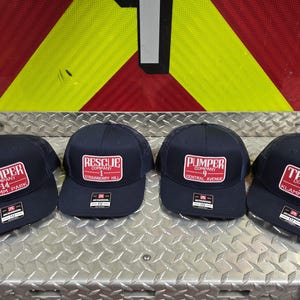 May include: Four navy blue baseball caps with red and white rectangular patches. The patches display text such as "Pumper Company 14 Klamm Park" and "Rescue Company 1 Strawberry Hill". The caps are arranged on a metal surface.