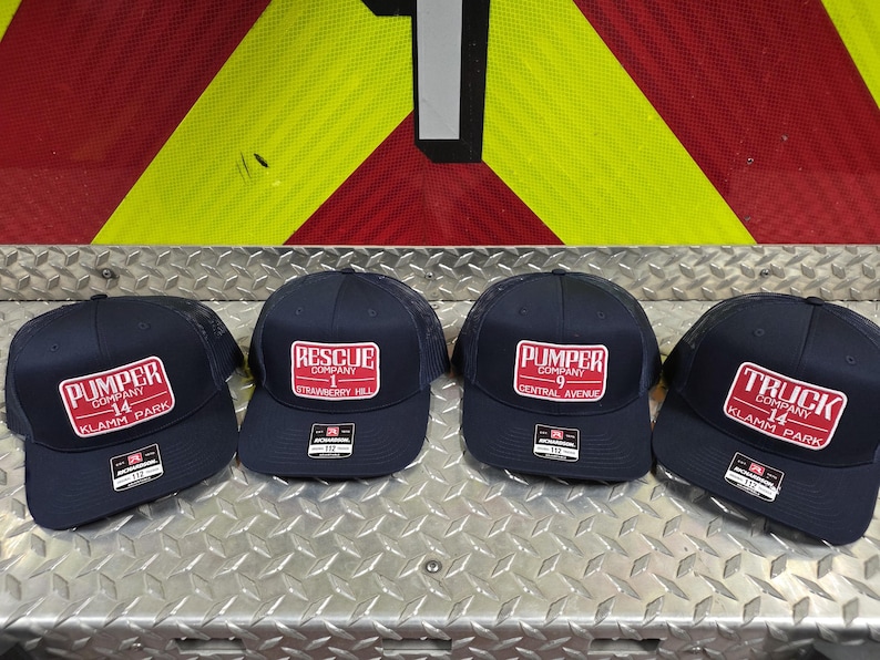 May include: Four navy blue baseball caps with red and white rectangular patches. The patches display text such as "Pumper Company 14 Klamm Park" and "Rescue Company 1 Strawberry Hill". The caps are arranged on a metal surface.