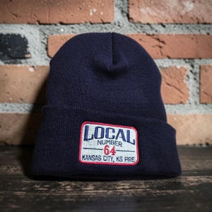 May include: A navy blue knit beanie featuring a rectangular embroidered patch on the cuff. The patch has a white background with a red border and blue text that reads LOCAL NUMBER 64 KANSAS CITY, KS FIRE.