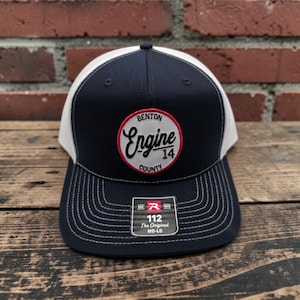 Pode incluir: Um boné trucker azul-marinho e branco com um emblema circular de borda vermelha e o texto Benton County Engine 14. O painel frontal exibe um adesivo preto com o texto 112 The Original MD-LG e um logotipo R vermelho na aba reta.