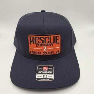 May include: Navy blue trucker hat with a mesh back and a curved brim. The front features a rectangular patch with the words "RESCUE COMPANY 2 FAIRFAX COUNTY, VA" in orange and white. The hat includes a Richardson tag.