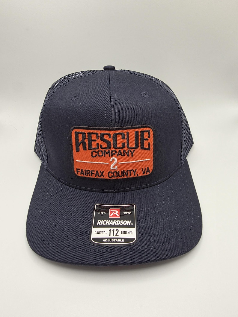 May include: Navy blue trucker hat with a mesh back and a curved brim. The front features a rectangular patch with the words "RESCUE COMPANY 2 FAIRFAX COUNTY, VA" in orange and white. The hat includes a Richardson tag.