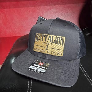 May include: A charcoal gray trucker hat with a mesh back. The front features a gold and black patch with the words "BATTALION CHIEF 1 KANSAS CITY, KS". The hat has a curved brim and an adjustable strap.