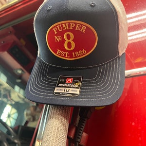 Op de afbeelding: Een marineblauwe en witte trucker-pet met een rode ovale patch waarop in gouden letters "Pumper No. 8 EST. 1886" staat. De pet heeft een verstelbare Richardson 112-riem.