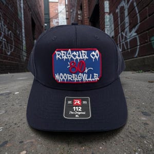 May include: A navy blue trucker hat with a mesh back and a rectangular patch on the front. The patch features a blue background with a red border and white text reading RESCUE CO 80 MOORESVILLE. A black sticker on the brim reads 112 The Original XL.