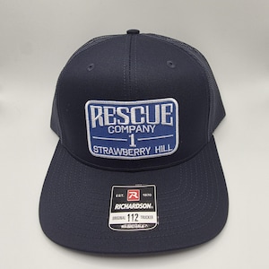 May include: Navy blue trucker hat with a mesh back and a curved brim. The front patch reads "RESCUE COMPANY 1 STRAWBERRY HILL" in white and blue. A Richardson tag is visible. Adjustable fit.
