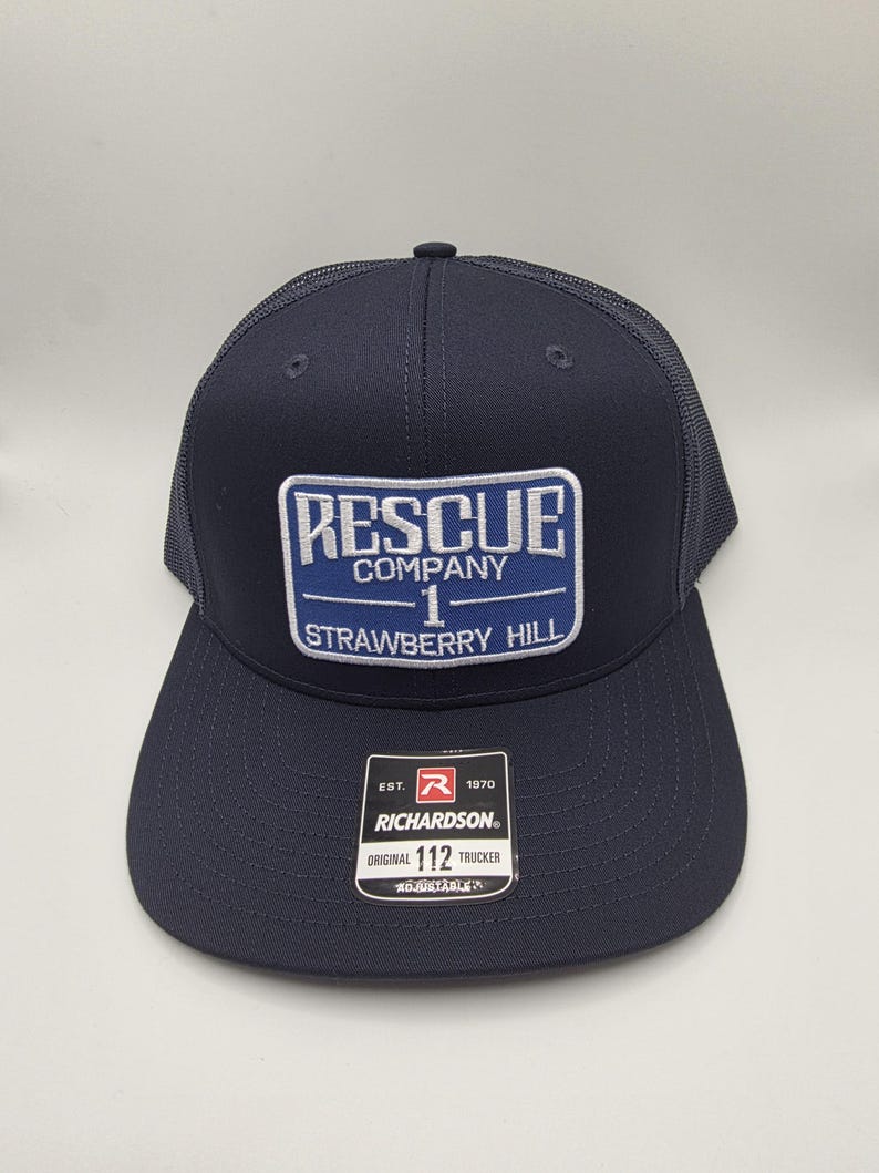May include: Navy blue trucker hat with a mesh back and a curved brim. The front patch reads "RESCUE COMPANY 1 STRAWBERRY HILL" in white and blue. A Richardson tag is visible. Adjustable fit.