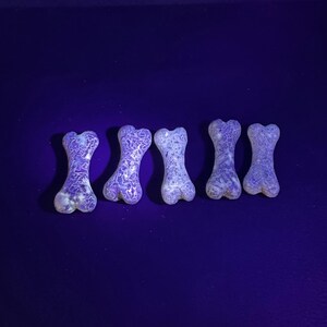 Puede incluir: Cinco objetos con forma de hueso dispuestos en una fila horizontal. Cada pieza presenta un patr&oacute;n jaspeado de color violeta y blanco brillante que fluoresce bajo luz ultravioleta, creando una textura intrincada y distintiva.