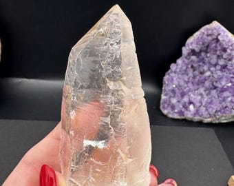 May include: A large, clear quartz crystal with a pointed tip. The crystal is translucent and has a slightly cloudy appearance. It is being held in a hand.