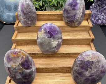 Puede incluir: Cinco piedras de amatista pulidas de diferentes tamaños y formas se exhiben en un soporte de madera escalonado. Las piedras exhiben una gama de tonos púrpuras con algunas áreas blancas y grises. Se ve follaje verde en el fondo.