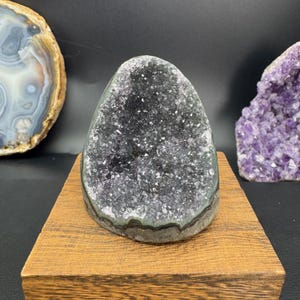 May include: A dark gray amethyst geode with sparkling crystal formations, displayed on a small wooden stand. The geode has a rounded, egg-like shape. Other geodes are visible in the background, including a sliced agate and a purple amethyst cluster.