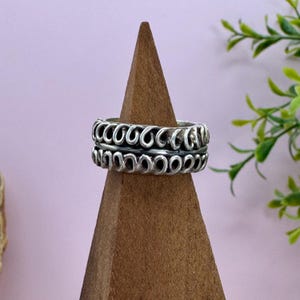 May include: A silver ring with a repeating open-loop design. The ring is displayed on a wooden ring holder against a soft pink background. The ring's design is a series of connected, curved shapes, creating a textured appearance.