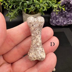 Puede incluir: Una peque&ntilde;a talla de piedra con forma de hueso que presenta un patr&oacute;n moteado e intrincado en tonos beige claro y marr&oacute;n tierra. La superficie muestra una textura compleja similar a una red en todo el objeto. La letra D est&aacute; impresa a la derecha.