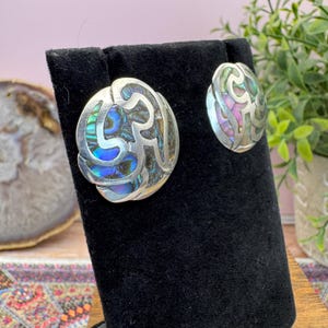 May include: A pair of silver-toned earrings featuring an intricate cutout metal design overlaid on iridescent abalone shell inlays. The earrings display swirling patterns with vibrant blue, green, and purple hues reflecting light across the shell surface.