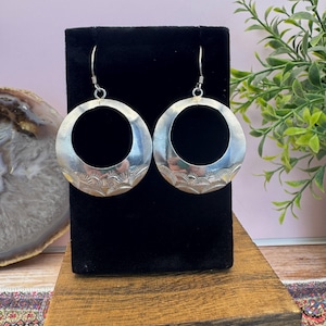 May include: A pair of silver-toned hoop earrings featuring a circular design with a polished upper section and a textured lower half displaying a repeating fan-like pattern. The earrings are suspended from simple silver-toned metal hooks.
