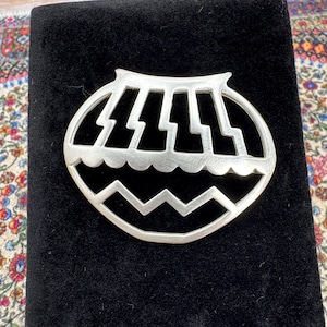May include: A silver-colored, openwork brooch in the shape of a stylized pot. The design features a series of vertical lines, a wavy line, and a zigzag pattern. The brooch is displayed on a black velvet surface.