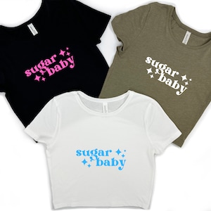 May include: Three cropped t-shirts with the text "sugar baby" in different colors. The first shirt is black with pink text, the second is olive green with white text, and the third is white with light blue text.