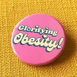 May include: A round, pink button with the words "Glorifying Obesity!" in white and teal lettering. The button has a glossy finish and is set against a yellow textured background.
