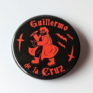 May include: Black button with red graphic of a figure holding a knife, with text "Guillermo de la Cruz" and bat and cross symbols. The design is a stylized illustration.
