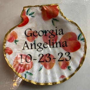 May include: A white seashell with a gold trim and a peach pattern. The shell has the text "Georgia Angelina 10-23-23" written on it.