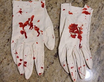 May include: A pair of white leather gloves with red blood splatters and streaks. The gloves are worn and have a distressed look.